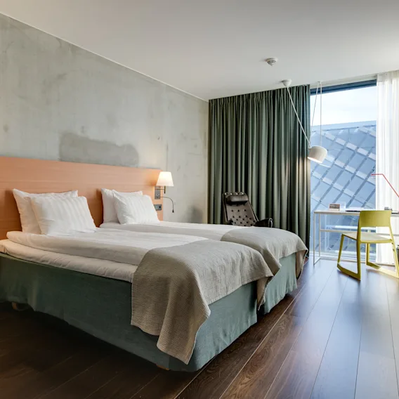 En neatly bäddad dubbelsäng står i ett modernt hotellrum, upplyst av vägglampor. I närheten står ett skrivbord och stol vid ett fönster med draperade gardiner.