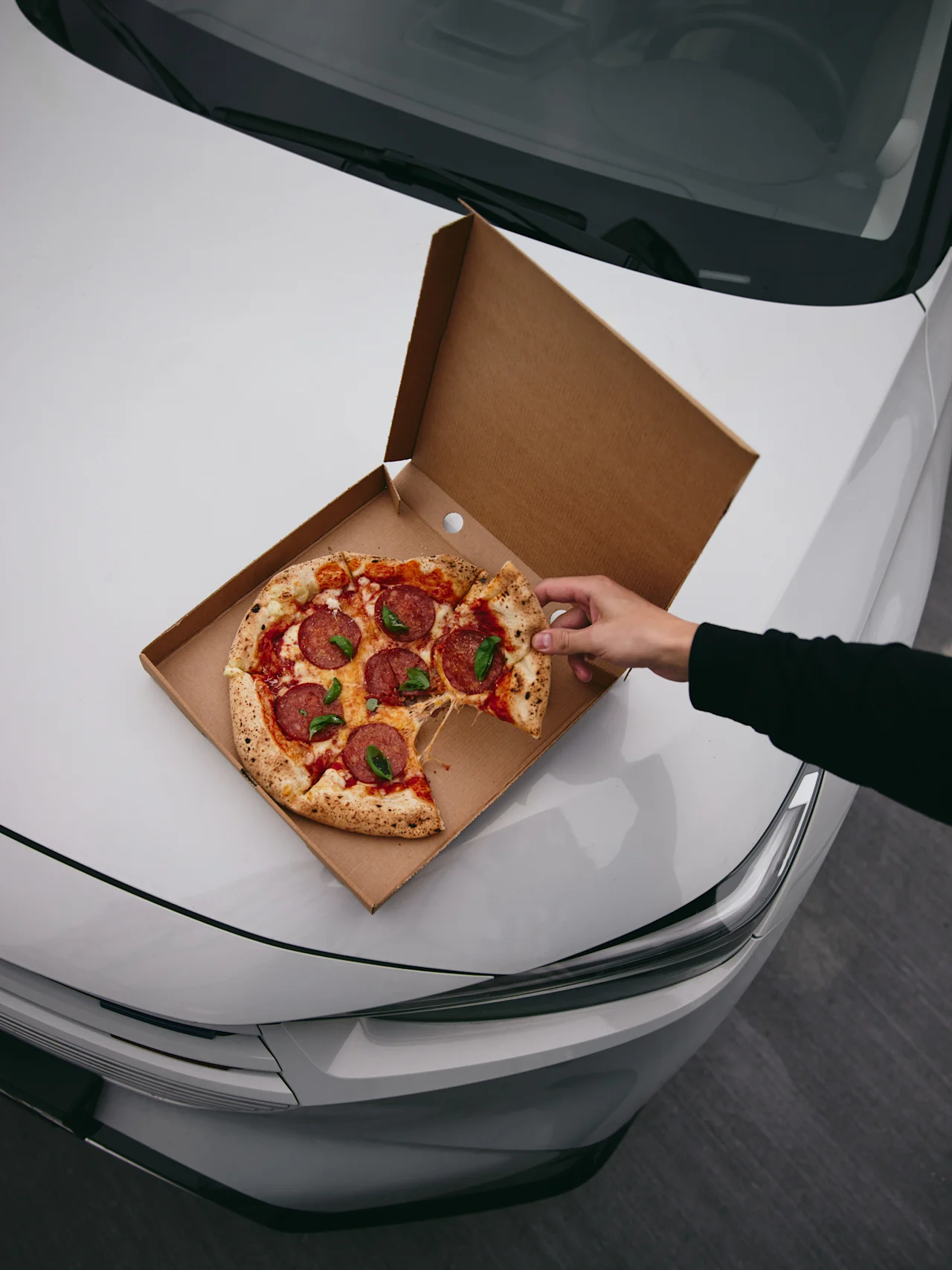 En hånd strekker seg etter et stykke pepperoni-pizza fra en eske som hviler på en bilpanser, et praktisk stoppestedmåltid på Hotell Falkenberg.