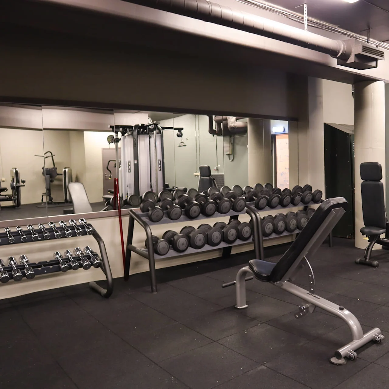 A gym with various weights and exercise machines on a rubber floor, industrial style with visible ductwork.