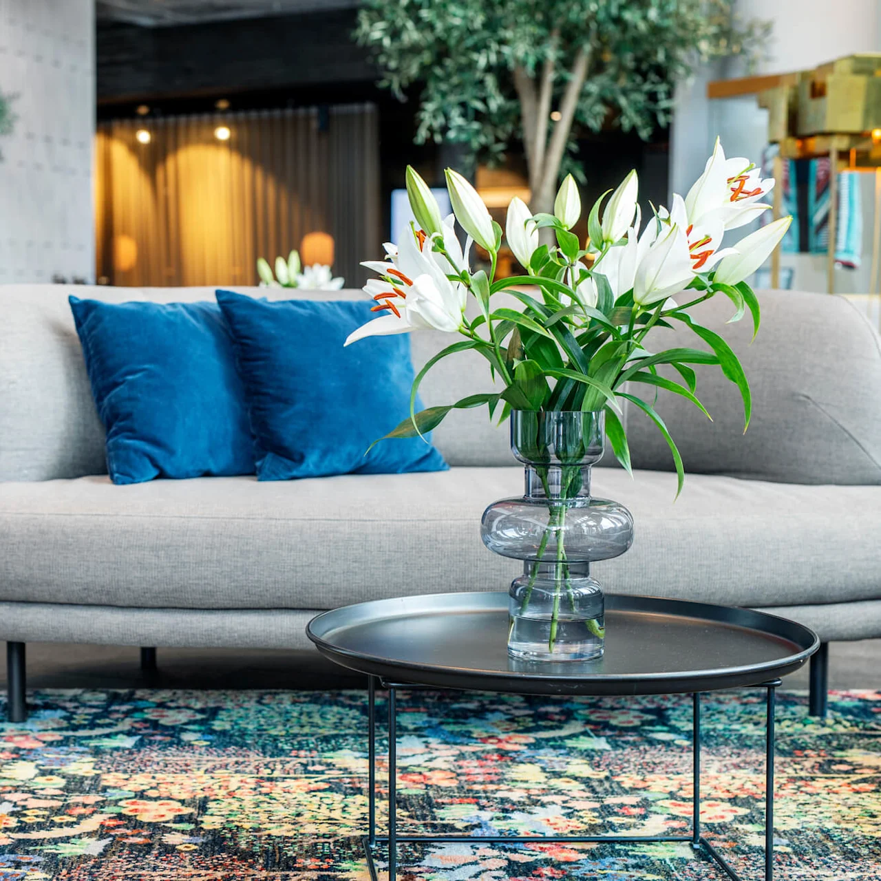 En grå sofa med blå puter er plassert i en moderne lounge. Et rundt bord har en glassvase med hvite liljer. Potted planter og myk belysning forbedrer inntrykket.