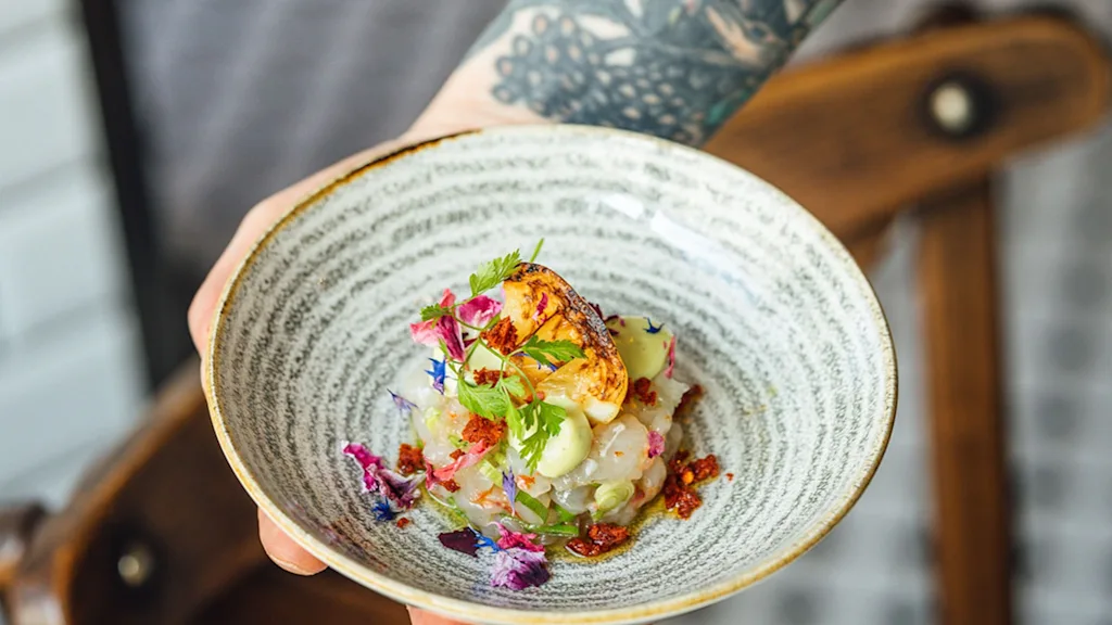 A tattooed arm holds a ceramic bowl with colorful, garnished seafood ceviche. The background features a blurred wooden chair and tiled wall.