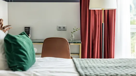 Ett mysigt hotellrum med en bekväm säng, grön kudde och mjuk stickad filt. Ett stilrent skrivbord finns under en lampa intill fönstret, inramat av djupa röda gardiner. En liten vas med en kvist ger en naturlig känsla.