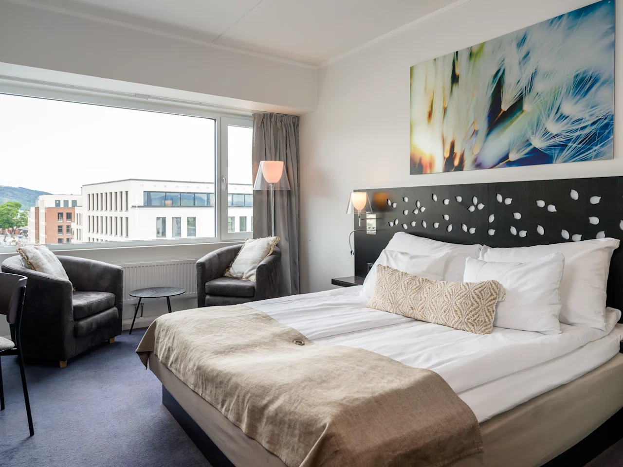 En prydligt bäddad säng med vita sängkläder och beige filt; omgiven av fåtöljer och bord nära ett stort fönster, konstverk ovanför sänggaveln i ett ljust, modernt hotellrum.