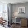 Lyst hotellrom med skrivebord, kontorstol, en behagelig skinnlenestol ved vinduet og et bilde på veggen.