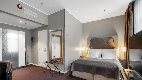 En pænt redt dobbeltseng med quiltet sengegavl og sengelamper er i et hotelværelse, omgivet af et vægspejl, garderobeskab og brunt tæppebelagt gulv.