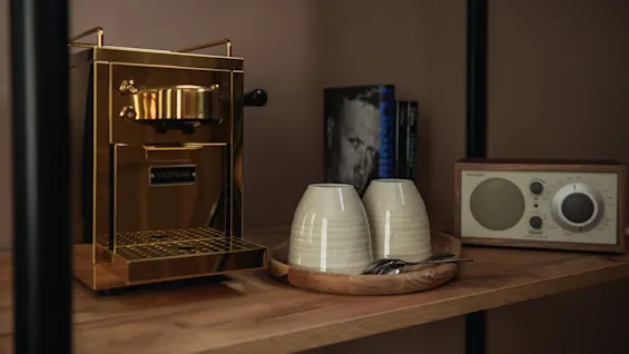 Golden coffee machine sits on a wooden shelf, with white mugs and spoons on a tray. A book and vintage radio are nearby. "SJÖSTRAND" is written on the machine.
