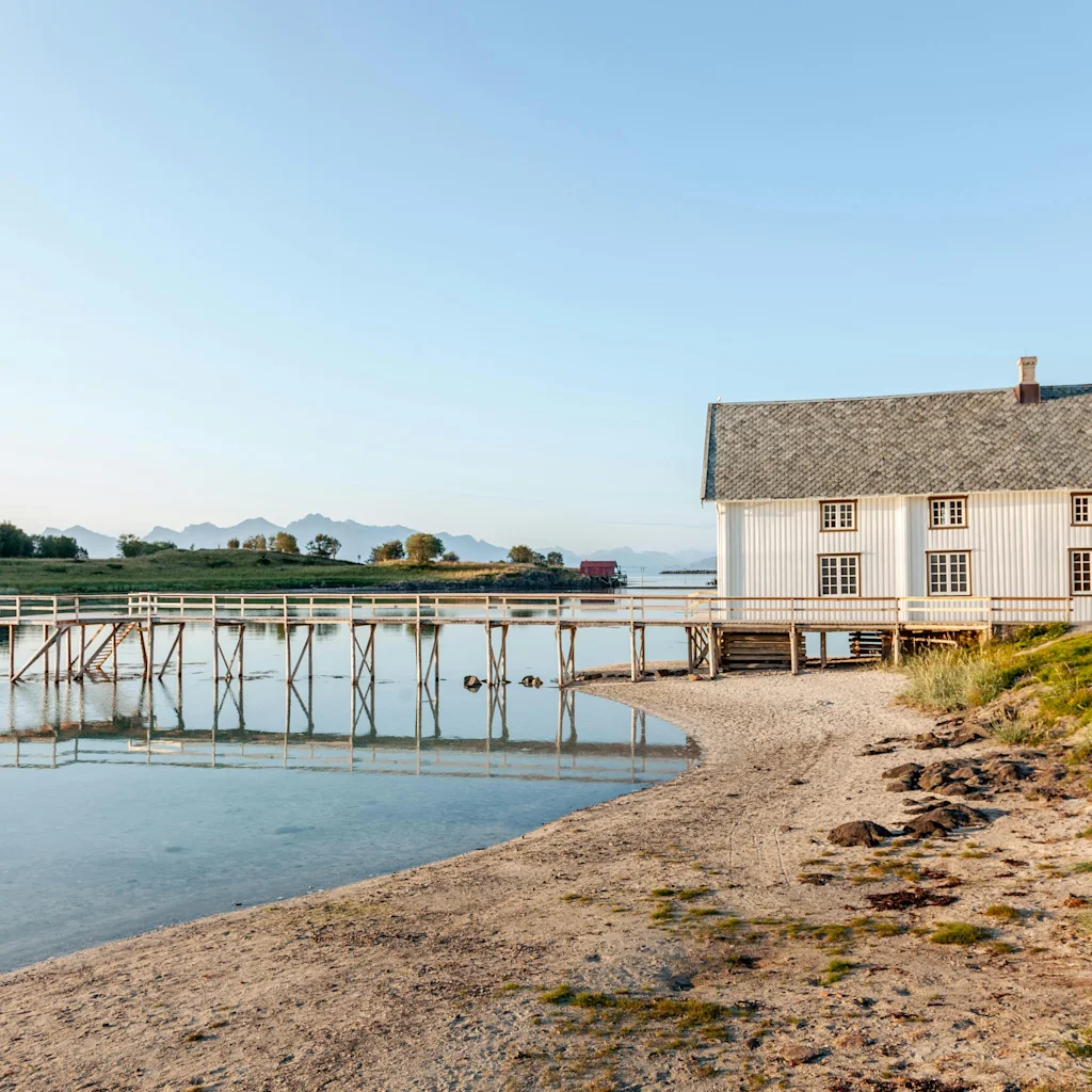 Ett vitt hus med grått tak står på en sandstrand vid lugnt vatten med en träbrygga. Berg i fjärran.