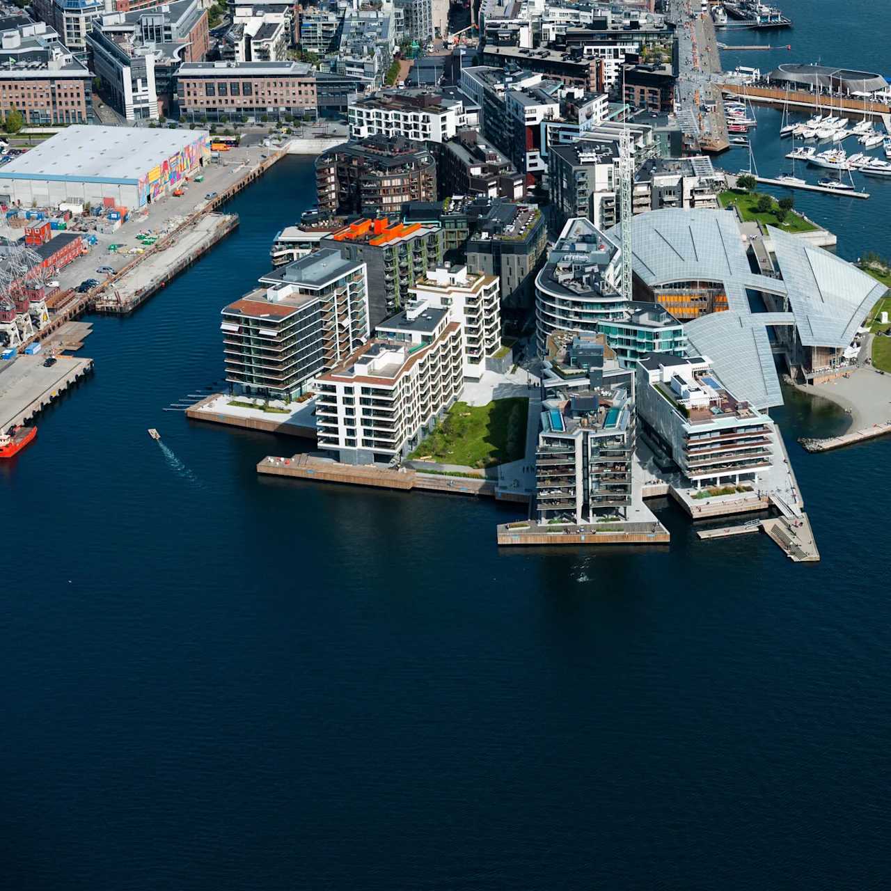 Luftfoto av et moderne havkompleks med vinklede bygninger og grøntområder; omgitt av en marina med hvite båter. Et høyt skip seiler på det rolige blå vannet i nærheten.