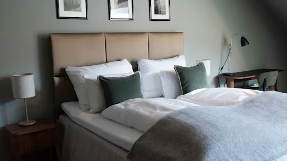 Et koselig hotellrom med en stor seng, myke puter og et varmt pledd. En lampe på nattbordet og et skrivebord med stol.