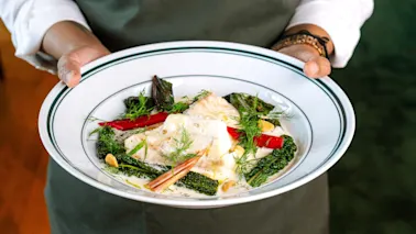 A person in a green apron holding a white plate with a delicious-looking meal of fish, green vegetables, and red peppers.