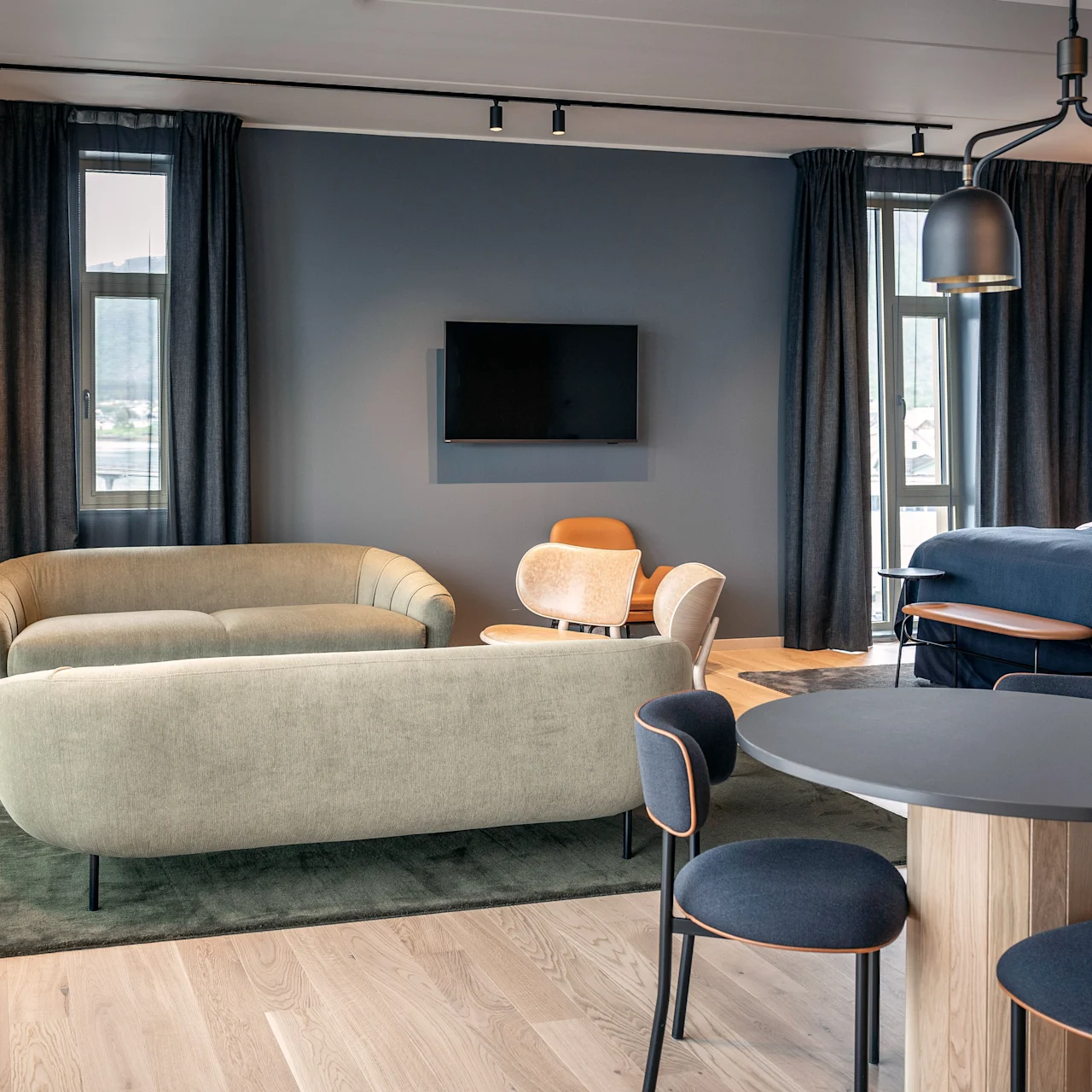 Et moderne opholdsstue har to sofaer på et tæppe, der vender mod et vægmonteret TV. Et rundt bord er omgivet af stole, med store vinduer og mørke gardiner, der giver naturligt lys.