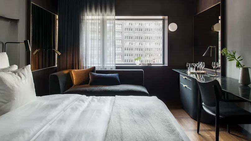 A modern hotel room features a neatly made bed, a dark couch with pillows, and a desk with a chair. A large window reveals an urban cityscape outside.