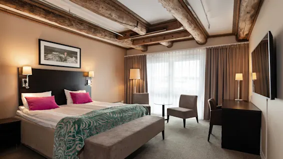 A neatly arranged hotel room features a large bed with pink pillows, a wooden desk, two chairs, and a patterned teal blanket, situated in a modern setting with wooden beam details.