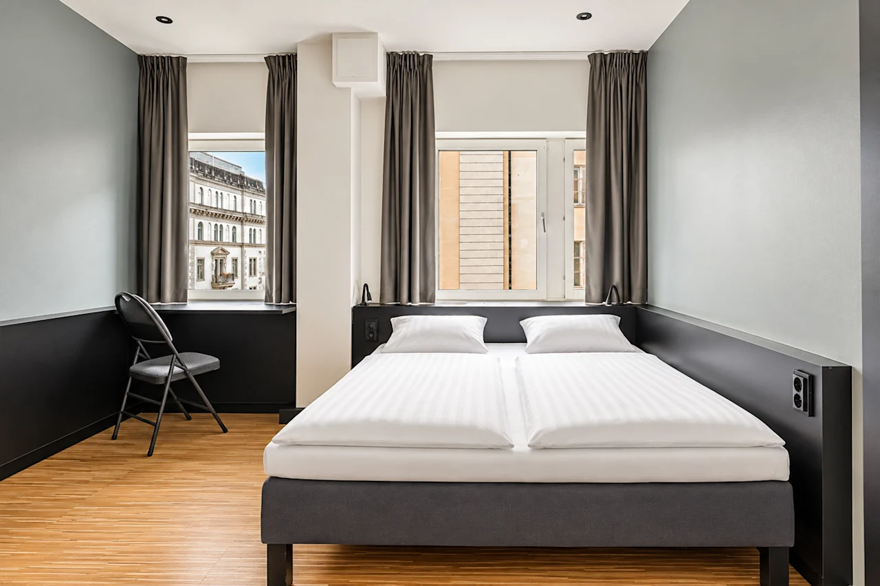Modern hotel room featuring a double bed, light walls, wooden flooring, and two windows letting in daylight.