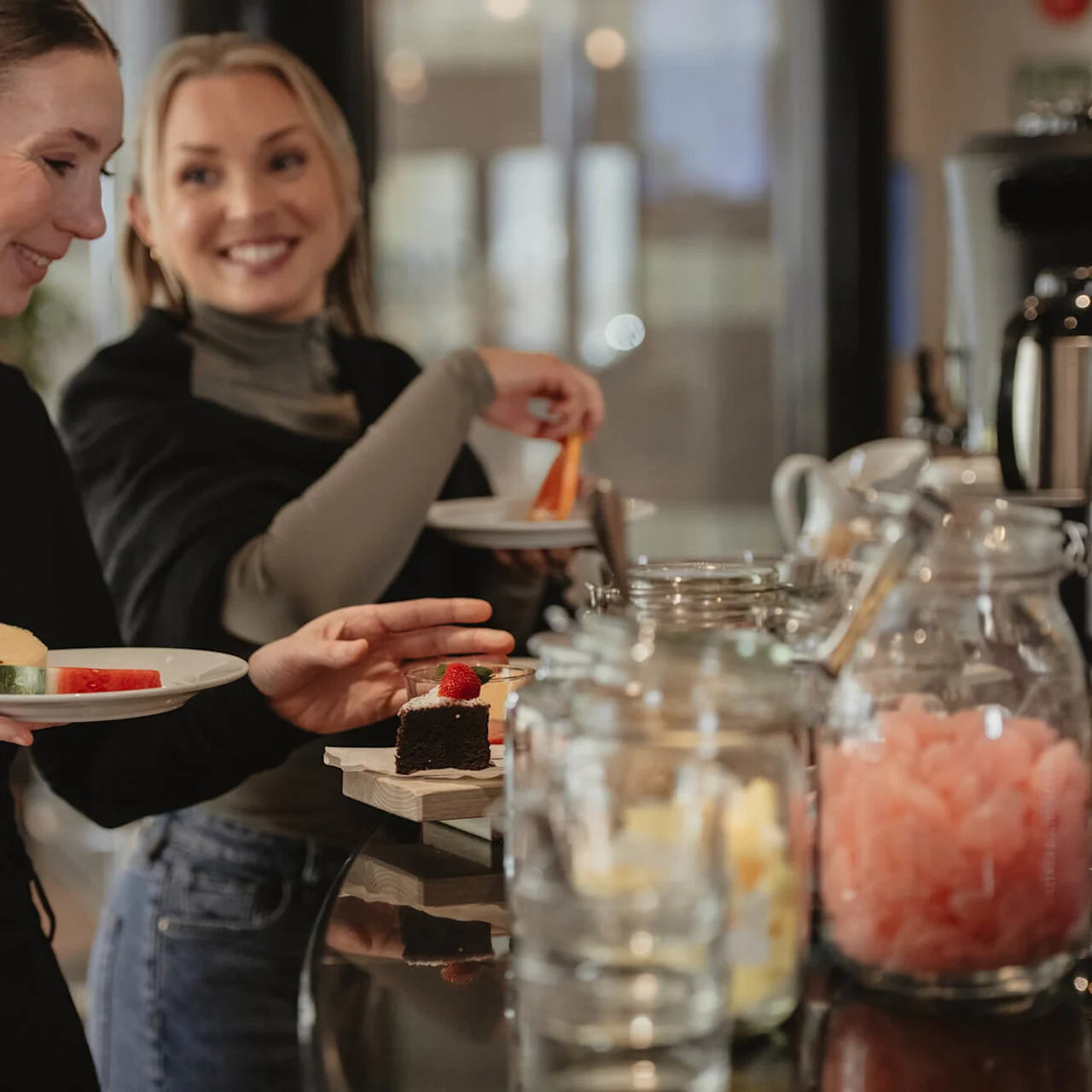 Two people smiling, holding plates with pastries and fruit, stand at a buffet table with jars in a cozy dining setting.