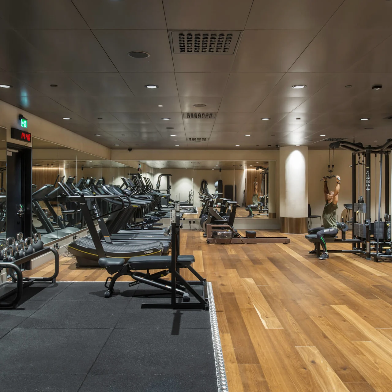 Løpemøller på rad i et godt opplyst treningsstudio, med en person som trener på en multigym-maskin. Parkettgulv og speil forsterker det romslige, organiserte miljøet. Digital klokke viser "14:40".