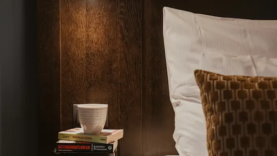 Cozy bedside at Home Hotel Kung Oscar with a warm lamp, books (SKAM, SKYMNINGSFLICKAN), and a cup, perfect for reading.