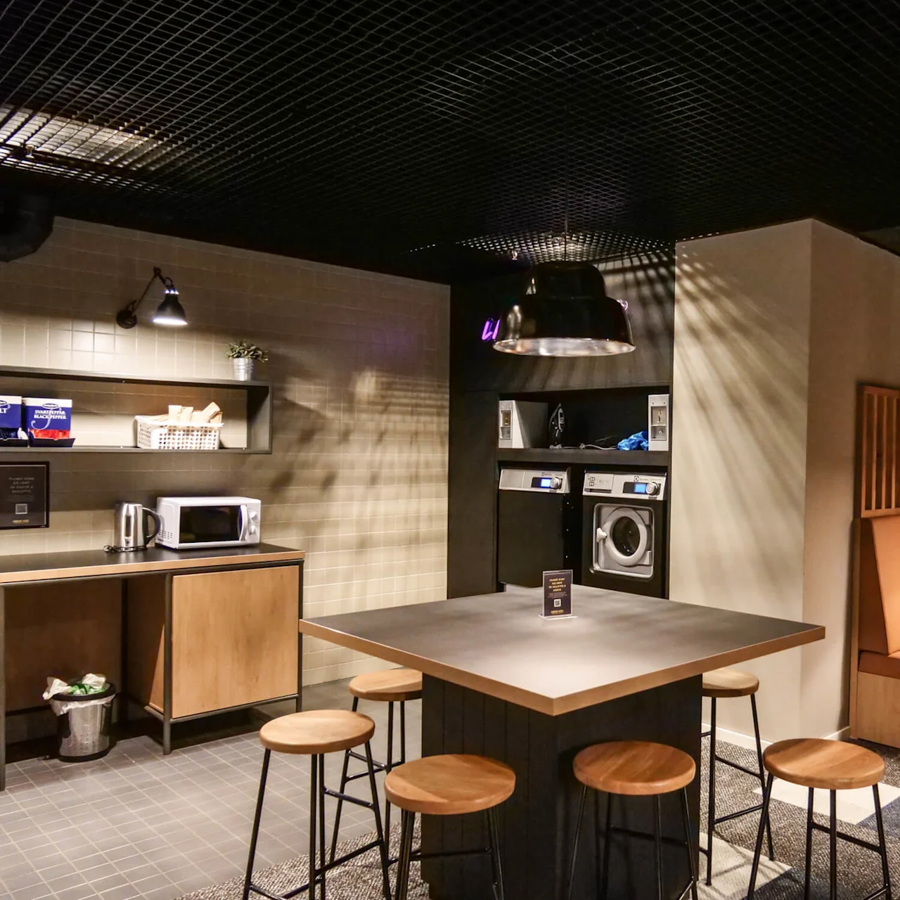 A kitchen features a central table with stools, surrounded by a microwave, kettle, washing machines, and seating area, creating a cozy, functional atmosphere. Text: "ret²" on the table sign.
