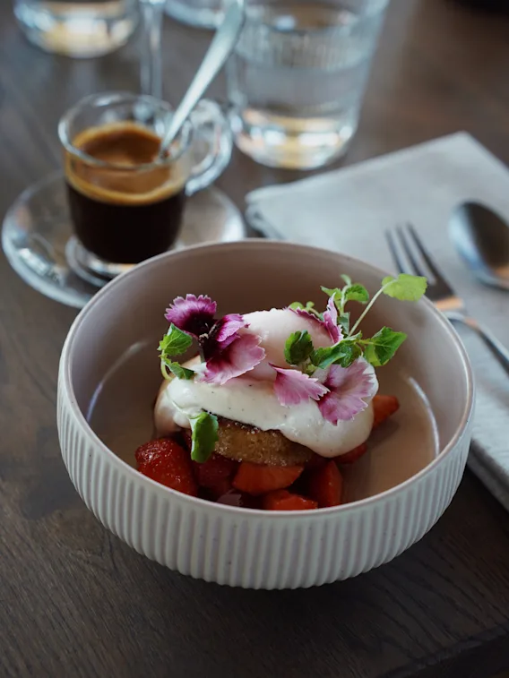 En vakkert anrettet dessert med jordbær, krem og spiselige blomster, servert sammen med en kopp kaffe og vann.