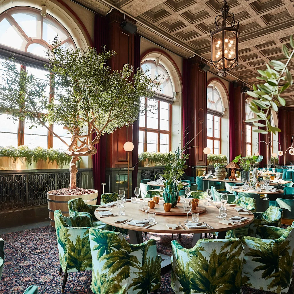Elegant spiseområde med store buede vinduer, frodige innendørstrær og stilige grønne sitteplasser med palmbladmønster.