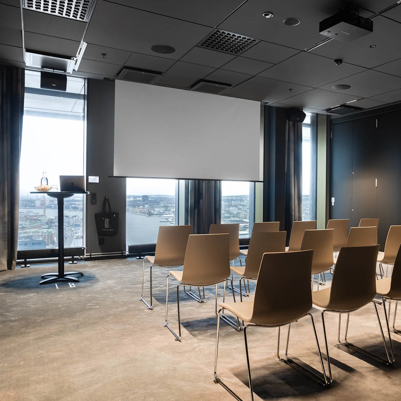 Rader av tomma stolar vända mot en stor projektorduk i ett modernt konferensrum med utsikt över staden genom höga fönster.