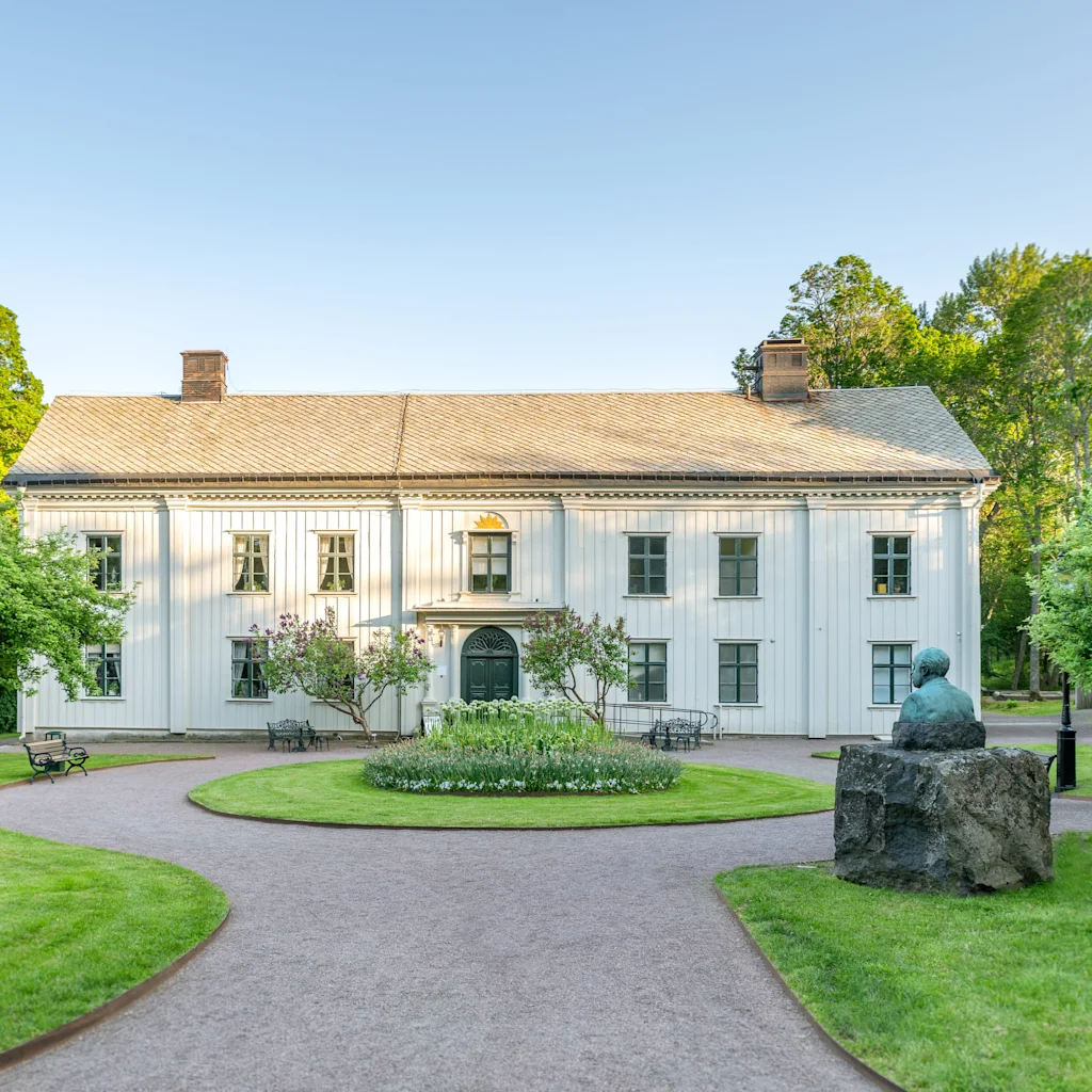 A grand white historic manor house surrounded by lush green lawns and trees under a clear blue sky