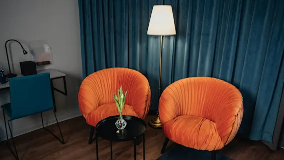 Two orange upholstered chairs sit beside a small black table with a vase of tulips, under a standing lamp, against blue curtains on a wooden floor. A desk with a blue chair is on the side.