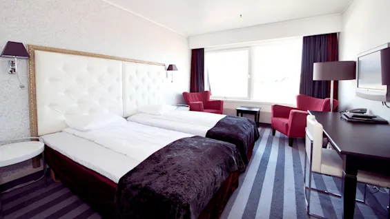 An elegantly decorated hotel room with two single beds, a red armchair, a desk, and a window.