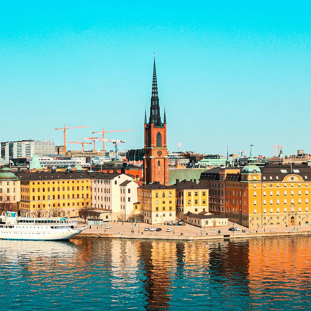 Stockholm cityscape with a church spire, colorful buildings, and a boat on the water under a clear blue sky.