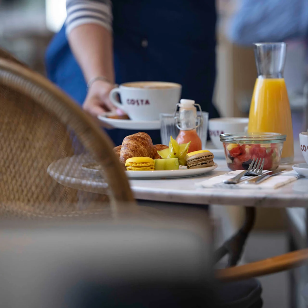 A table features a breakfast spread with pastries, macarons, and fruit. Orange juice and a cappuccino with the brand "COSTA" on the cup are present, served by a person.