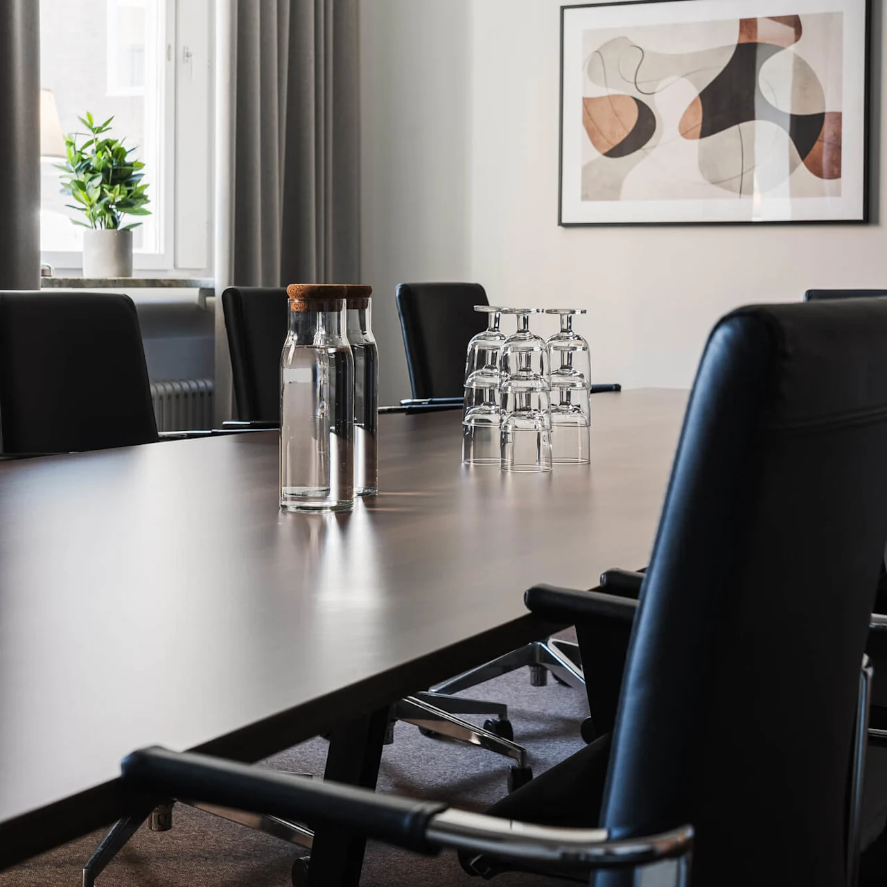 Konferensbord med vattenflaskor och uppochnedvända glas; omgiven av svarta stolar. En växt står i fönstret bredvid abstrakt konstverk på krämfärgad vägg.