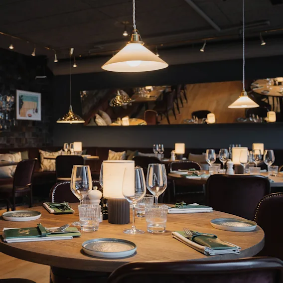 Round dining table set with wine glasses, plates, and napkins; surrounded by cushioned chairs and illuminated by pendant lights in an elegant dimly-lit restaurant with art-adorned walls.