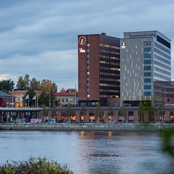 En klockförsedd hotellbyggnad står upplyst vid en lugn flod, omgiven av andra byggnader och träd under en molnig himmel. Text: "Dorsia" och "Elite Hotel."