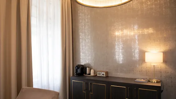 Et hyggeligt værelse med en kaffestation på et mørkt skab, lyst vindue med gardiner og varm belysning.