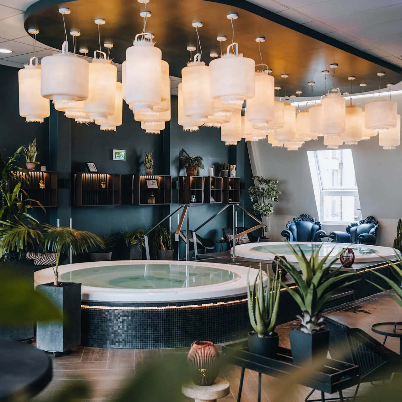 A round jacuzzi rests under a ceiling adorned with lanterns, surrounded by green plants and modern decor. The setting appears to be a luxurious, well-lit spa room.