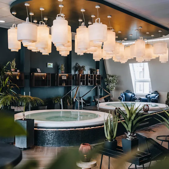 En rund jacuzzi ligger under ett tak dekorerat med lyktor, omgiven av gröna växter och modern inredning. Miljön verkar vara ett lyxigt, välbelyst spa-rum.