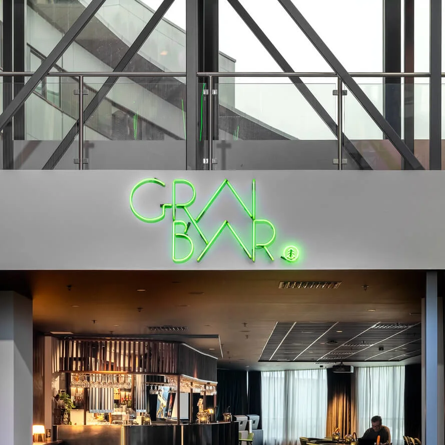 A bar with modern decor features a neon sign reading "GRØN BYAR." Inside, tables and chairs are arranged, with a person seated, creating an inviting and relaxed atmosphere.