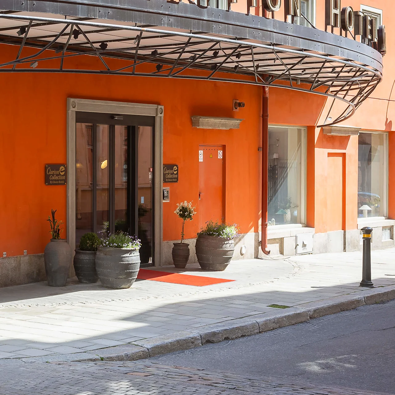 En orange byggnad med ett böjt metalltak över en trottoar med stora fönster, krukväxter och en röd matta vid ingången.