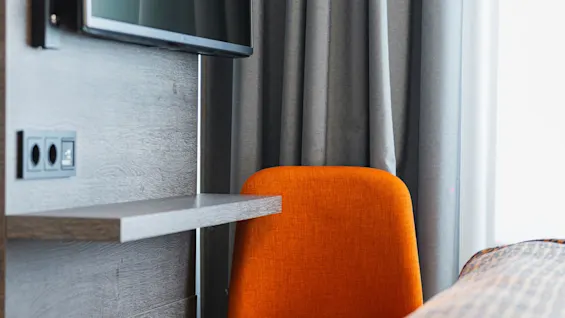 En orange stol står vid en väggmonterad TV och hylla. I närheten är grå gardiner delvis öppna, och en säng med ett orange-mönstrat täcke finns i förgrunden.