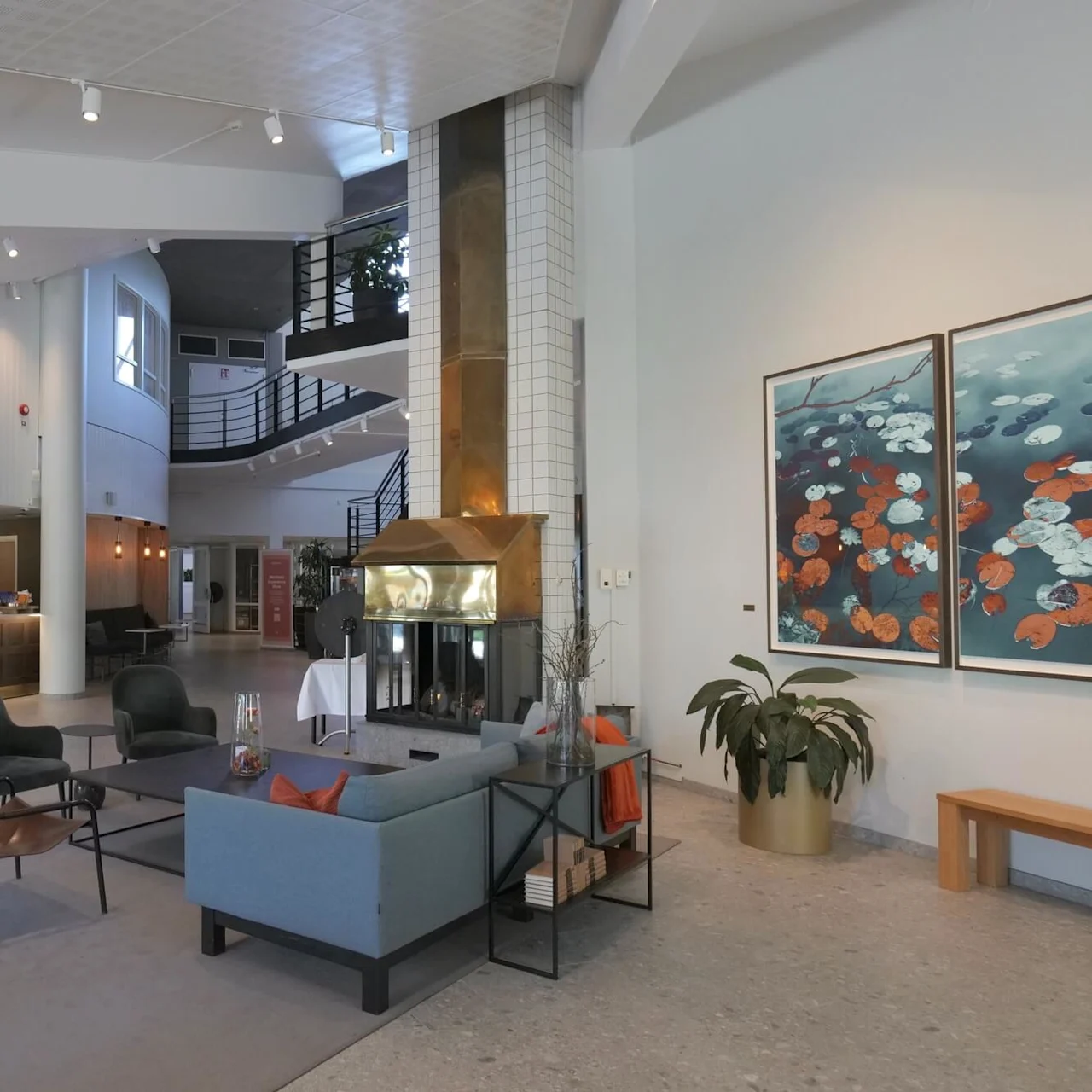 Sofagruppe omgir et glassbord, vendt mot en moderne peis. Bak, en buet trapp fremhever den romslige lobbyatmosfæren, komplett med resepsjonsdisk og dekorative malerier.