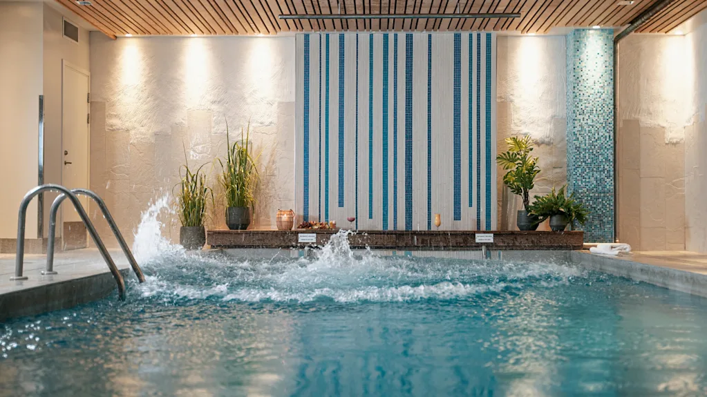 Poolvatten plaskar energiskt mot en dekorerad inomhusspa-vägg med blå kakel och växter. Varma ljus och träpaneltak ger en avslappnande atmosfär.