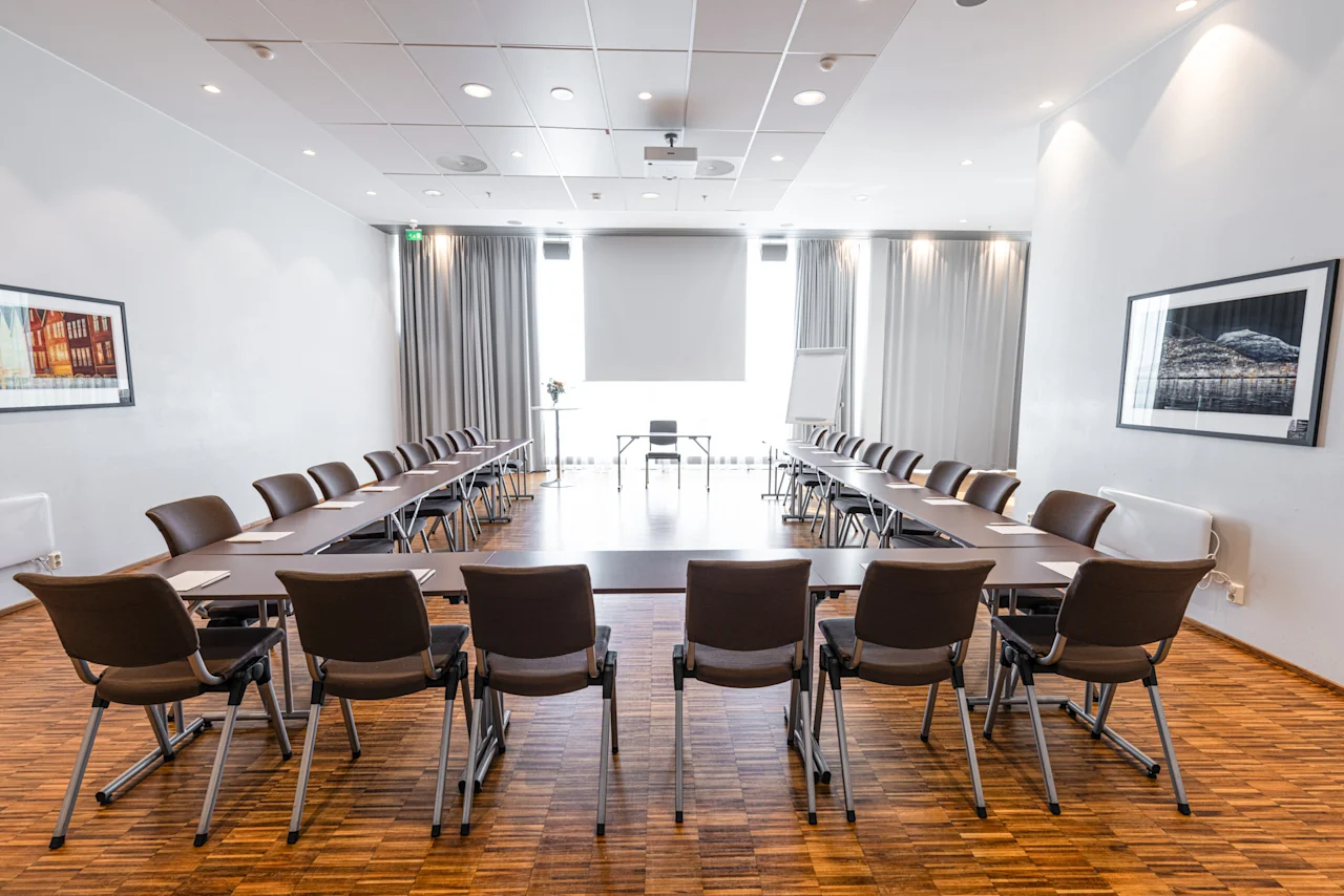 U-muotoinen konferenssipöytä, jossa on mustia tuoleja, on asetettu valoisaan huoneeseen, jossa on puulattia ja kehystettyjä taideteoksia seinillä.