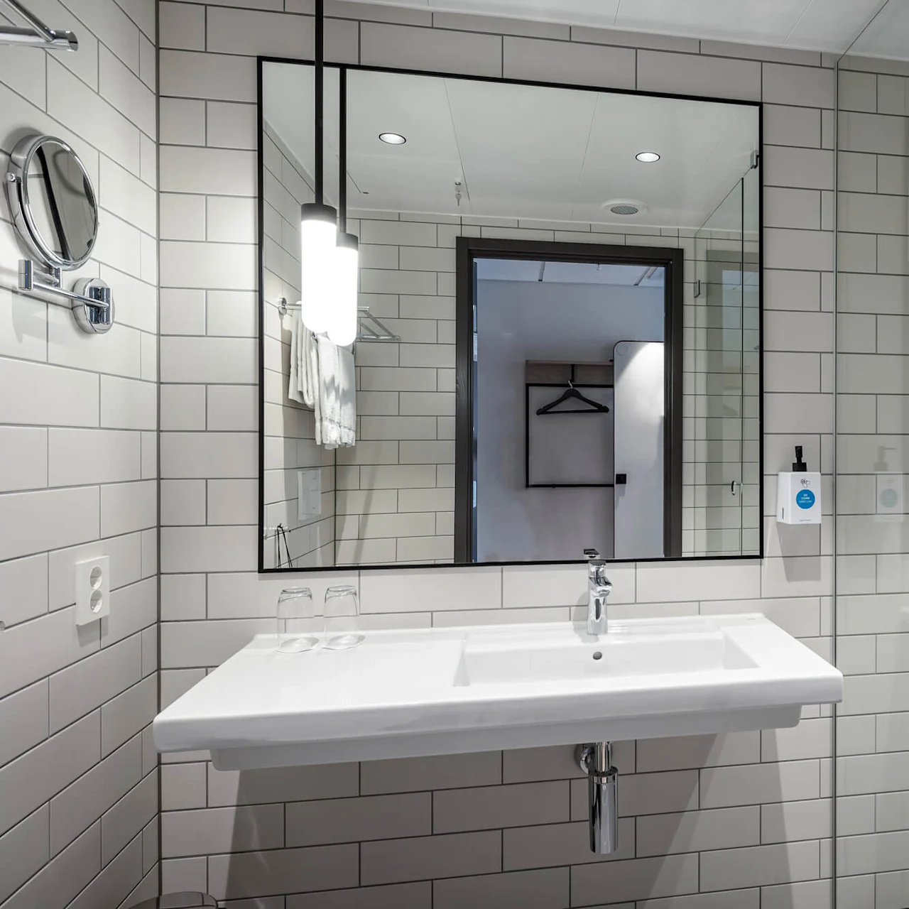 Vask reflekteres i et badeværelse spejl, omgivet af hvide flisevægge; en glasbruser med regnbruserhoved er synlig til højre, og toiletartikler er monteret på væggen.
