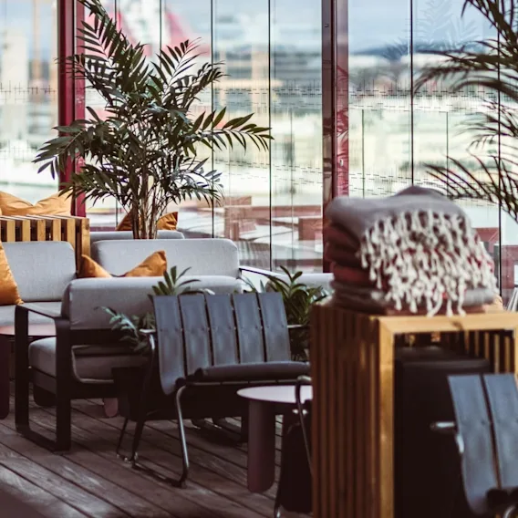 Komfortable sitteplasser med grå puter og oransje puter er ordnet utendørs på et trebord dekket med potteplanter, ved siden av et glassveggområde med byutsikt.