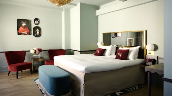 A large bed covered with white linens and red pillows sits in a room with light walls, red chairs, and a round table. Unique animal-themed artwork decorates the walls.