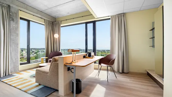 A bright, modern room with large windows overlooking a city, featuring a comfortable sofa and a functional desk area.