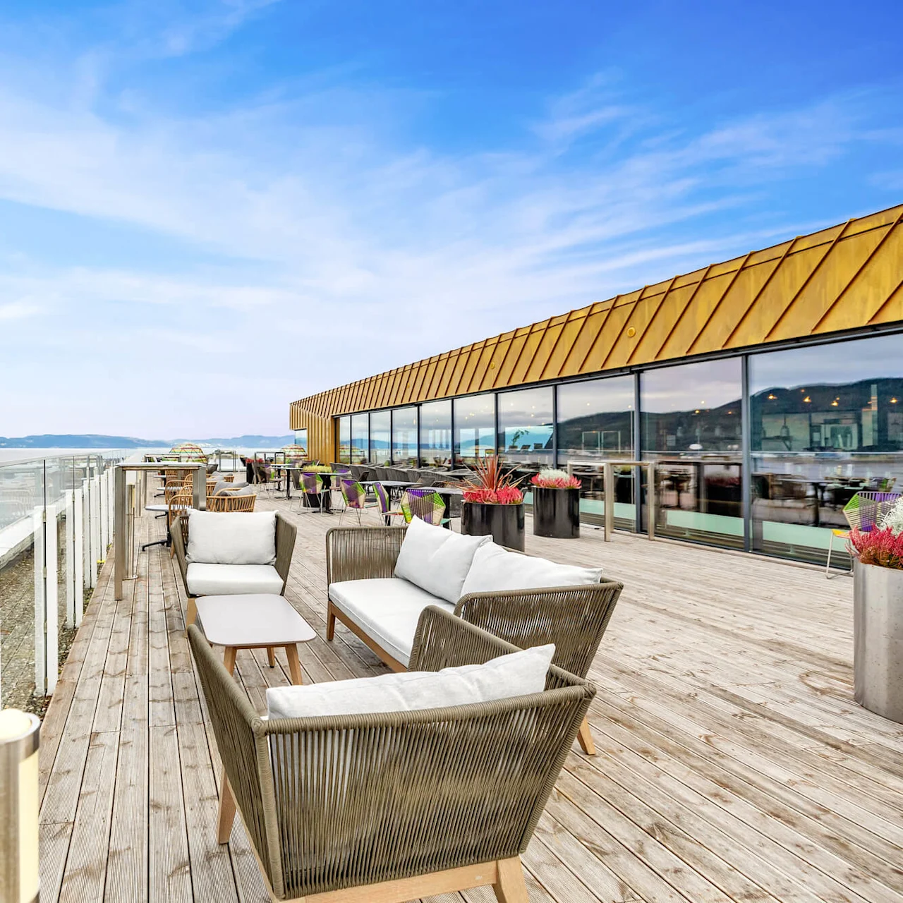 Terrace with wicker furniture arranged near potted plants on wooden decking, overlooking a waterfront view. Glass railing offers open scenery, while large windows reflect the sky.