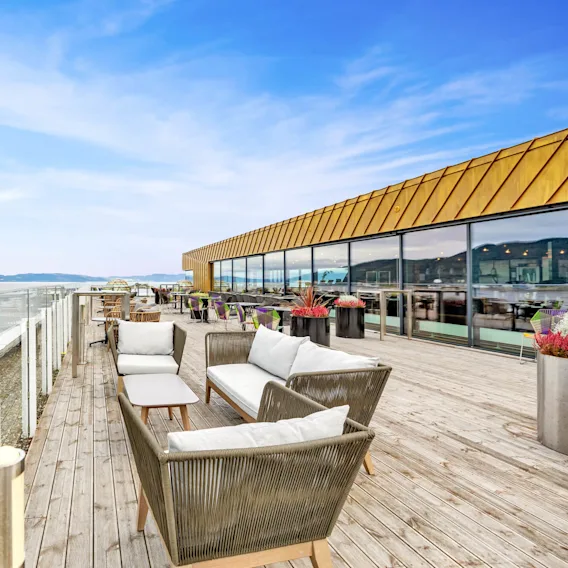Terrasse med kurvmøbler plassert nær potteplanter på tregulv, med utsikt over vannet. Glassrekkverk gir åpen utsikt, mens store vinduer reflekterer himmelen.