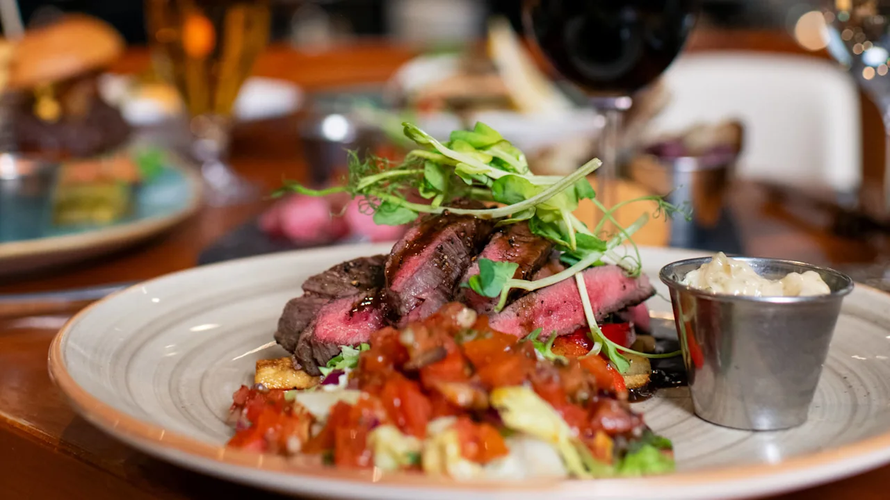 En tallerken med skiver af oksekød toppet med salat serveres med ristede grøntsager og en sovsekop. To glas, et med rødvin og et med øl, står i nærheden i en restaurationsomgivelse.