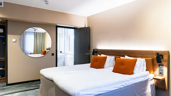 En pænt redt dobbeltseng med orange puder står i et moderne hotelværelse. Et rundt spejl reflekterer dele af værelset, og en åben dør afslører et kig ind i badeværelset.
